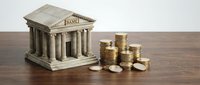 Miniature bank building model with classical columns next to stacks of golden coins on wooden surface representing financial services and savings.