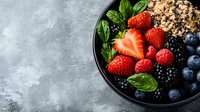 Fresh mixed berries and granola bowl with strawberries, blueberries, blackberries, raspberries and basil leaves on dark plate against grey concrete background.