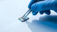 Gloved hand holding a micro SD card with tweezers on a light surface, showcasing precision in electronics handling and data storage technology.