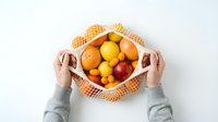 Hands holding mesh bag filled with fresh oranges, lemons, and apples on white background. Overhead view for healthy eating, grocery shopping, and organic produce concepts.