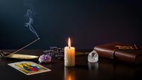 Mystical fortune telling setup with burning candle, tarot cards, amethyst crystal, incense stick and leather journal on dark surface for spiritual guidance.
