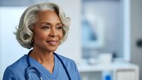Confident mature African American female doctor with gray hair wearing blue scrubs and stethoscope in modern medical office setting.