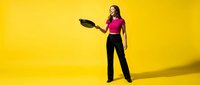 Young Hispanic woman chef in pink top holding black frying pan against bright yellow background for culinary marketing and cooking advertisements.