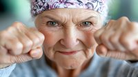 Senior Caucasian woman with floral headscarf making playful fists and smiling mischievously at camera, expressing joy and vitality in aging with positive attitude.