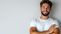 Confident young Hispanic man with beard wearing white t-shirt, standing with arms crossed against light wall background, expressing positive friendly attitude.