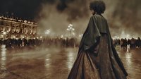 Young African American woman in vintage cloak standing alone in foggy night square, watching crowd in distance against illuminated historic building backdrop.