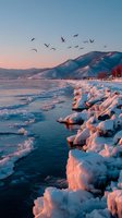 Frozen shoreline with ice formations along mountain lake at sunset, birds flying over winter landscape bathed in pink light.