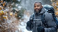 African American man with beard wearing winter jacket smiling during snowfall, carrying backpack outdoors. Active lifestyle and winter adventure concept.