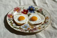 Two sunny side up fried eggs on toast served on a vintage floral porcelain plate, placed on a white linen cloth in natural light.