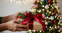 Hands holding wrapped gift box with red ribbon bow and pine sprig decoration against blurred Christmas tree lights background for holiday celebrations.