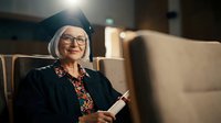 Happy senior Caucasian woman in graduation cap and gown smiling in auditorium. Academic achievement, lifelong learning, university ceremony concept.