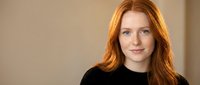 Young redhead woman with freckles smiling at camera in casual black sweater against neutral background for professional headshot and portrait use.