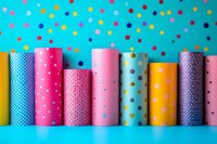 Colorful polka dot wrapping paper rolls arranged in row against bright turquoise background with confetti pattern, festive craft supplies for gift packaging.