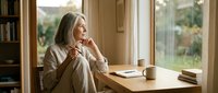 Mature woman with long gray hair sitting at wooden desk by window, wearing beige cardigan, thoughtfully resting chin on hand with coffee cup and books nearby, natural daylight interior setting.