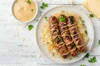 Grilled bratwurst sausages served on sauerkraut with creamy sauce drizzle and fresh parsley garnish, perfect for oktoberfest menu, restaurant promotion or food blog content.