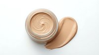 Creamy beige foundation makeup in glass jar with smooth swirl texture and smear sample, isolated on white background for cosmetic design projects.
