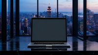Laptop with blank screen on desk overlooking city skyline at dusk, suitable for website mockups, business presentations, and corporate advertising.