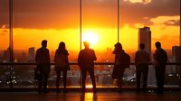 Silhouettes of diverse young professionals watching dramatic orange sunset over city skyline from modern office building or observation deck. Urban lifestyle scene.