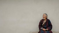 Senior Asian woman with gray hair and glasses wearing a dark burgundy cardigan over a patterned shirt, seated against a neutral beige wall, smiling warmly while looking to the side, suitable for healthcare, wellness, aging, and lifestyle editorial use.