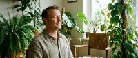 Middle-aged Caucasian man relaxing in bright living room surrounded by lush green houseplants creating peaceful indoor garden atmosphere for wellness content.