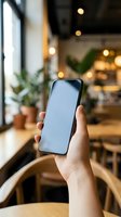 Hand holding smartphone with blank white screen in cozy cafe interior, mockup for app or website display, copy space on screen.