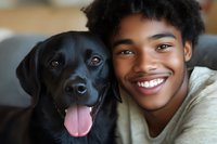 Young African American woman with natural curly hair smiling while cuddling with black Labrador retriever dog on couch, expressing joy and companionship.