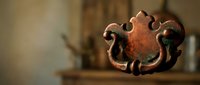 Antique copper drawer pull with ornate baroque design and aged patina, set against a warm blurred rustic interior background.