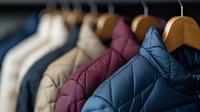 Winter jackets in blue, burgundy and beige hanging on wooden hangers in retail store closet, showing quilted pattern texture and modern outerwear fashion display.