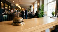 Vintage brass service bell on wooden hotel reception desk with blurred restaurant interior background for hospitality and customer service concepts.