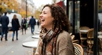 Young Hispanic woman with curly hair laughing joyfully at outdoor cafe terrace during autumn day. Genuine happiness and positive emotions in urban setting.