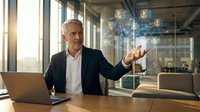 Mature businessman in suit interacting with holographic network interface at modern office desk with laptop during golden hour sunlight.