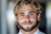 Young Caucasian male with blonde wavy hair, blue eyes, and trimmed beard wearing white shirt smiles warmly at camera in natural lighting headshot portrait.