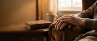 Elderly person's wrinkled hands resting on a chair arm, warm golden light, vintage book and mug on wooden table, cozy home interior.