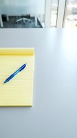 Blue ballpoint pen resting on yellow lined notepad at modern office desk with windows and blurred workspace background, ready for writing and note-taking.