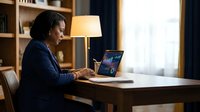 Professional African American businesswoman working on laptop at wooden desk in modern office with warm lighting and bookshelf background.