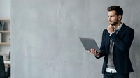 Caucasian male professional in dark blazer holding laptop, hand to chin in thoughtful pose against concrete wall, suitable for business and corporate concepts.