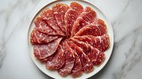 Thinly sliced cured meat arranged in circular pattern on white ceramic plate against marble background, showcasing delicate marbling and rich red color.