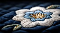 Detailed macro shot of elegant embroidered flower with white and blue petals on dark fabric, featuring delicate stitching and golden accents. Traditional needlework artistry.