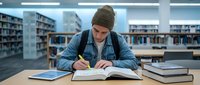 Young male student in beanie hat studying at library table with books and tablet, taking notes in academic environment for education and learning concepts.