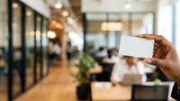 Cropped hand holding blank white business card in modern office environment with blurred colleagues and glass partitions in background.