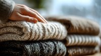 Woman's hand touching soft knitted sweaters stack, cozy winter clothing collection, warm wool garments pile, textile comfort and fashion concept.