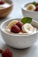 Creamy chocolate smoothie bowl topped with fresh raspberries, sliced banana and mint leaves in speckled ceramic bowl on white background, selective focus.