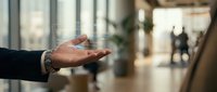 Mature male hand extended in welcoming gesture inside modern office lobby with blurred colleagues and glass architecture in background, representing hospitality, business greeting, and professional environment.