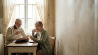 Senior Caucasian couple enjoying morning coffee together at kitchen table by window, reading newspaper in cozy home interior for retirement lifestyle.