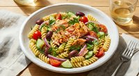 Colorful fusilli pasta salad with cherry tomatoes, olives, herbs and cheese in white bowl on wooden table with wine glass for Mediterranean cuisine.