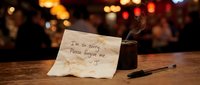 Handwritten apology note on a bar counter reading I'm so sorry, please forgive me, with a smoking candle and pen in a moody setting.