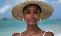 Young African American woman wearing straw sun hat and hoop earrings smiling at beach, turquoise ocean and blue sky background, summer vacation lifestyle portrait.