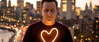 Middle-aged Caucasian man wearing glowing neon heart symbol on chest standing against blurred city skyline at sunset for love and connection concepts.