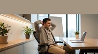 Young professional man relaxing at modern office desk with laptop, stretching back in chair during work break in bright contemporary workspace.