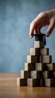 Hand placing graduation cap on wooden blocks pyramid representing education achievement and career advancement concept for academic success.